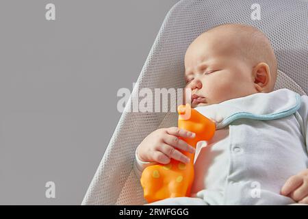 Le bébé dort avec une girafe en caoutchouc jouet orange dans une chaise longue sur un fond gris avec de l'espace pour copier Banque D'Images