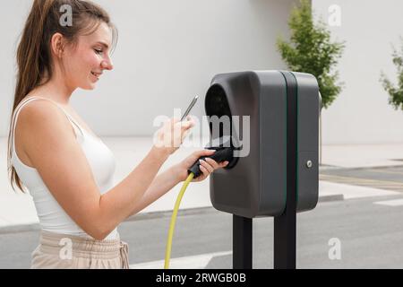 Fille connectant sa voiture électrique et une station de charge avec un chargeur de câble et appuyant sur un bouton de démarrage pour activer une session de charge Banque D'Images