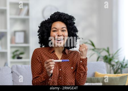 Portrait rapproché d'une jeune femme afro-américaine assise sur le canapé à la maison, tenant un test rapide unique dans ses mains et heureuse d'apprendre un résultat positif pour la grossesse. Banque D'Images