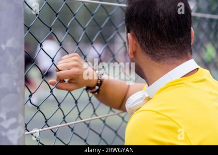 homme avec écouteurs regarde les joueurs de football sur l'arène urbaine, regardant à travers la grille de clôture métallique. Banque D'Images