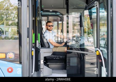 Jeune adulte homme chauffeur de navette de ville assis dans la cabine du conducteur en attendant les passagers à l'arrêt de bus. Banque D'Images