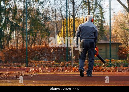 Homme aux cheveux gris portant des cache-oreilles à l'aide d'un souffleur de feuilles portable pour nettoyer la zone clôturée du parc. Vue arrière de l'homme senior en salopette bleu foncé, soufflant les feuilles du terrain de jeu. Concept de travail. Banque D'Images