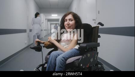 Femme avec l'atrophie musculaire spinale en fauteuil roulant motorisé au milieu du couloir de la clinique sourit et regarde la caméra. Le personnel médical marche dans le couloir de l'hôpital ou de l'établissement médical en arrière-plan. Banque D'Images