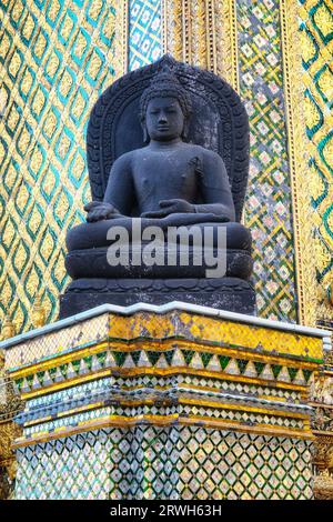 Une statue de Bouddha sereine. La statue sombre, assise sur une base, est placée sur un fond animé de motifs complexes en or, vert forêt et bleu ciel Banque D'Images