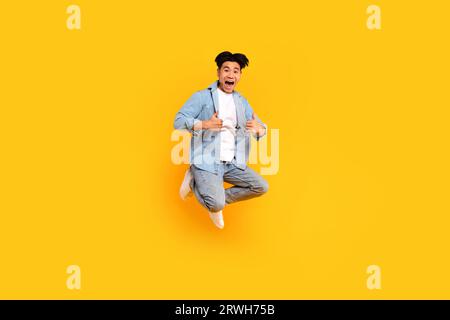 Homme asiatique excité en tenue élégante montrant les pouces tout en sautant sur fond de studio jaune, recommander quelque chose Banque D'Images