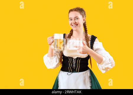 Jeune femme dans les vêtements traditionnels allemands pointant vers la bière sur fond jaune Banque D'Images