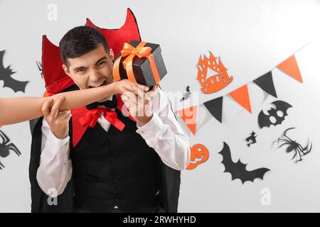 Jeune homme habillé pour Halloween comme vampire mordant la main avec cadeau sur fond clair Banque D'Images