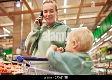 Jeune mère avec son bébé mignon dans le chariot se promène dans le supermarché et parle au téléphone. Belle maman et petite fille blonde shopping Banque D'Images