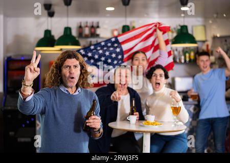Groupe d'amis supporters acclamant pour l'équipe préférée avec drapeau des États-Unis tout en regardant match à la télévision dans le pub Banque D'Images
