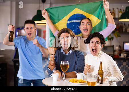 Compagnie de jeunes fans émotifs pour adultes soutenant l'équipe sportive du Brésil avec drapeau d'état tout en se reposant avec la bière au bar Banque D'Images