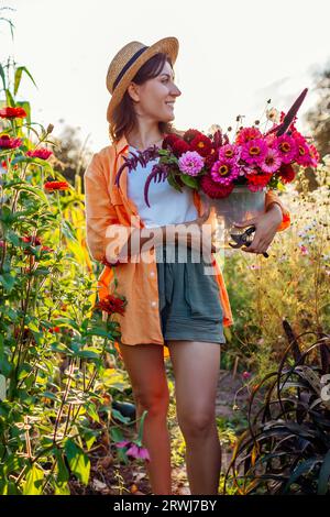 Portrait de femme fermière cueillant des fleurs dans le seau dans le jardin d'été au coucher du soleil. Récolte de fleurs coupées de zinnias amarante dahlias. Banque D'Images