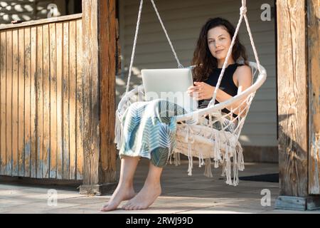 une jeune femme se repose sur une chaise de balançoire macrame avec ordinateur portable sur la terrasse de la maison de campagne