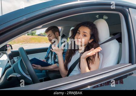 Jeune femme assise sur la place du conducteur et regardant la caméra avec confusion pendant que l'instructeur remplit le rapport d'essai routier. Test de conduite, conducteur Banque D'Images