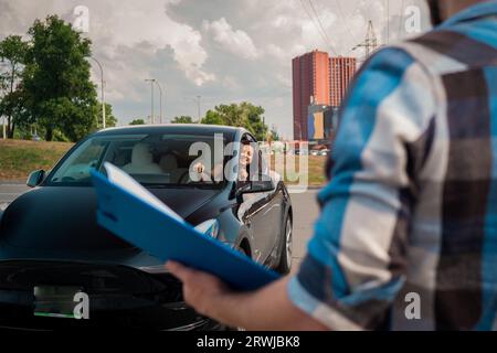 Femme souriante apprenant à conduire et regardant par la fenêtre de la voiture, regardant l'instructeur avec le rapport d'essai. Test de conduite, cours de conducteur, concept d'examen Banque D'Images