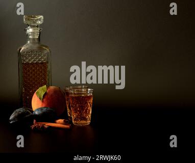 Liqueur de prune-pêche dans une carafe en cristal et deux verres avec cannelle, anis et fruits mûrs sur fond noir. Gros plan. Banque D'Images