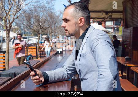 Homme d'affaires caucasien adulte en costume élégant, inquiet assis à l'extérieur du restaurant à midi en utilisant le téléphone et en pensant. Banque D'Images