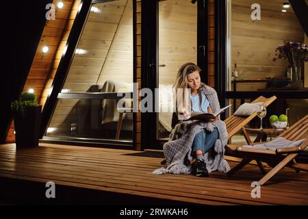 Comme le soir tombe, une femme s'enroule dans un plaid et lit un livre sur la terrasse de la cabane en rondins Banque D'Images