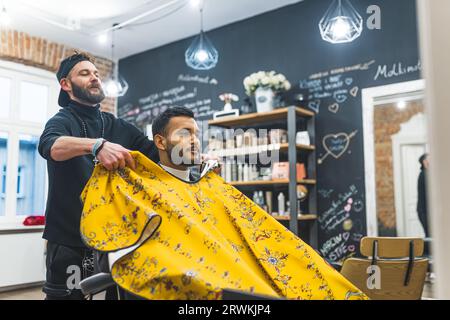 Barber mettant une cape imprimée jaune sur son client indien masculin avant une coupe de cheveux. Photo de haute qualité Banque D'Images