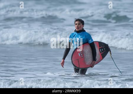 Un jeune surfeur mâle transportant son surf et marchant hors de la mer après avoir conté à la compétition de surf RIP Curl Grom Search au Fistral au ne Banque D'Images