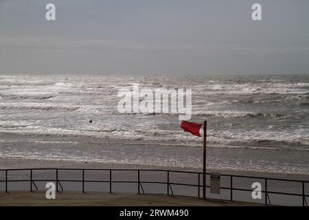 Deux personnes au loin dans une mer agitée, ignorant un avertissement de drapeau rouge de danger, mer sombre reflétant la dernière lumière du soleil Banque D'Images