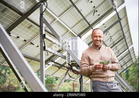 Investisseur calculant la somme qu'il a gagné en investissant. Homme heureux souriant à cause du profit qu'il a obtenu après avoir investi dans des innovations alternatives. Homme enthousiaste souriant et regardant droit la caméra. Banque D'Images