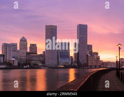 Canary Wharf et la Tamise à l'aube avec un ciel orange doré derrière les immeubles de bureaux modernes de grande hauteur. Mur de premier plan, chemin et lampadaires Banque D'Images