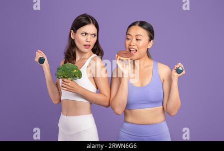 Femme asiatique mangeant un beignet tandis que son amie tenant le brocoli et la regardant, debout sur fond violet Banque D'Images