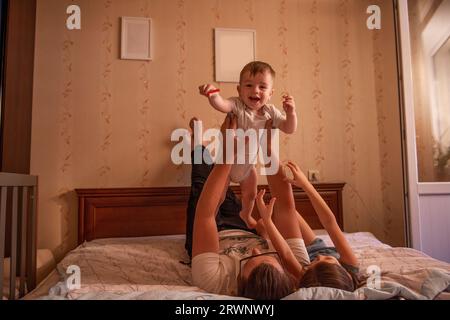 Le père diversifié dans les lunettes jette bébé fils sur le lit, la fille aînée s'amuse à proximité. La famille fous autour et joue le week-end. Da du père Banque D'Images