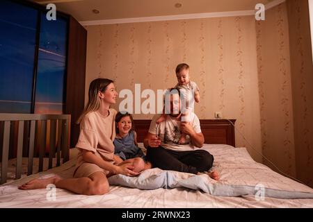 Portrait drôle de famille joyeuse diverse sur le lit le matin. Mère élégante embrasse sa fille, père avec des lunettes tient le petit fils sur les épaules. Parents Banque D'Images