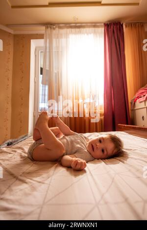 Body bébé d'un an repose sur le dos, pendant les jambes sur le lit le matin dans les rayons du soleil. L'enfant est drôle, souriant, jouant de bonne humeur. Entretien A. Banque D'Images