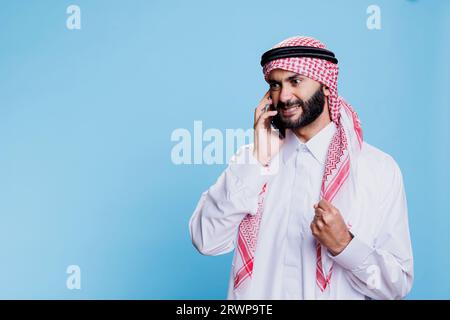 Homme musulman furieux portant des vêtements traditionnels se mettant en colère tout en parlant sur le téléphone portable. arabe agressif debout avec le poing serré et l'expression en colère tout en répondant à un appel sur smartphone Banque D'Images