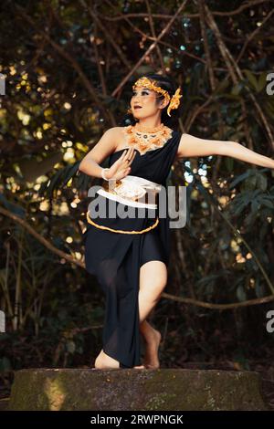 Femme balinaise dansant sur le sommet du rocher tout en portant un costume noir et une couronne dorée sur sa tête à l'intérieur de la jungle Banque D'Images