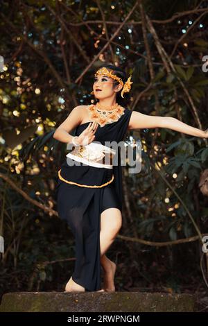 Femme balinaise dansant sur le sommet du rocher tout en portant un costume noir et une couronne dorée sur sa tête à l'intérieur de la jungle Banque D'Images