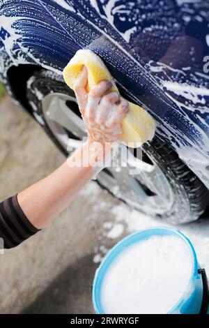 Homme lavant la voiture bleue avec une éponge jaune et un seau d'eau savonneuse Banque D'Images