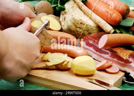 Pelez les pommes de terre pour la soupe de pommes de terre Disposé devant des légumes frais, du bacon et des saucisses Banque D'Images