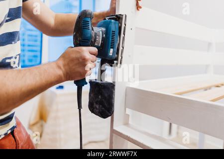 l'homme méconnaissable frotte le lit d'enfant en bois avec une ponceuse électrique ou un broyeur en enlevant la vieille peinture. Image avec mise au point sélective Banque D'Images