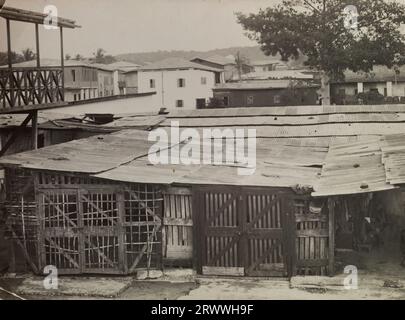 Vue d'un bidonville à Accra, montrant des toits en tôle ondulée fixés au-dessus des portes et des planches de bois fixées ensemble de manière lâche. Il y a des gens à l'intérieur des habitations, et des arbres et des bâtiments plus permanents derrière. Légende manuscrite ultérieure : Shanty Town Accra. Banque D'Images