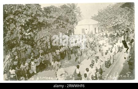 Une rue bondée à Winneba comme une procession du Vendredi Saint fait son chemin à travers une route bordée d'arbres, de bâtiments et de spectateurs. Deux longues files de femmes habillées intelligemment défilent dans la rue avec un orchestre qui se produit au milieu d'elles et des gens qui suivent derrière le groupe portant des drapeaux. Sous-titré en négatif : procession indigène...Gold Coast. Banque D'Images