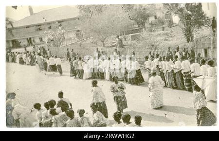 Deux longues files de femmes se rassemblent dans la rue, prêtes à commencer une procession. Tous sont habillés avec élégance dans des chemisiers blancs et des jupes à motifs. Il y a des bâtiments visibles derrière eux. La légende d'une image d'accompagnement décrit l'événement comme une procession du Vendredi Saint à Winneba. Banque D'Images