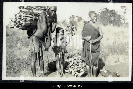 Un groupe de deux femmes Kikuyu avec un enfant, toutes vêtues de châles traditionnels et de bijoux, se tient au bord d'une route. Une femme porte un paquet de bois sur son dos et l'autre repose à ses côtés. L'enfant tient un bouquet de fleurs sauvages. Légende originale du manuscrit : femmes Kikuyu portant du bois. Banque D'Images