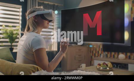 Femme dans le casque VR choisit le film ou l'émission de télévision à regarder à la maison. Hologramme futuriste 3D montre l'interface de l'application de service de streaming et des widgets dans le menu utilisateur. Animation VFX. Concept de style de vie. Banque D'Images