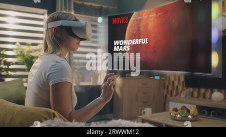 Femme dans le casque VR choisit le film ou l'émission de télévision à regarder à la maison. Hologramme futuriste 3D montre l'interface de l'application de service de streaming et des widgets dans le menu utilisateur. Animation VFX. Concept de style de vie. Banque D'Images