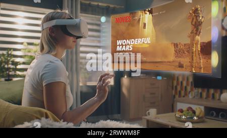 Femme dans le casque VR choisit le film ou l'émission de télévision à regarder à la maison. Hologramme futuriste 3D montre l'interface de l'application de service de streaming et des widgets dans le menu utilisateur. Animation VFX. Concept de style de vie. Banque D'Images