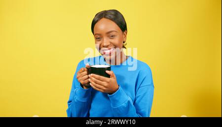 Contented Black Woman sirote une boisson chaude de mug, fond de studio jaune Banque D'Images