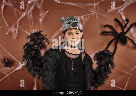 Joyeuse fille preteen posant dans la tenue noire Halloween et masque de loup sur fond brun Banque D'Images