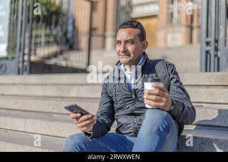 Portrait d'un homme latino assis sur un escalier avec un téléphone portable et une tasse de café jetable. Banque D'Images