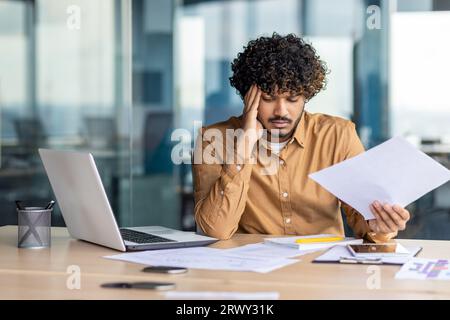 Financier contrarié regardant les ventes et le tableau des revenus de l'entreprise, homme d'affaires au sein du bureau déçu par les résultats et les réalisations des ventes, homme hispanique assis à table avec des documents et un ordinateur portable. Banque D'Images