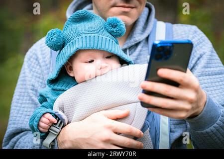 Père et nouveau-né dans une écharpe dans la forêt d'automne, homme avec téléphone portable. Famille heureuse. Banque D'Images