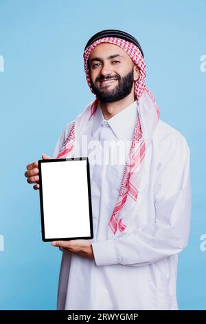 Homme musulman souriant montrant tablette numérique avec écran blanc vide et regardant la caméra avec une expression joyeuse. Heureux annonceur arabe tenant le portrait blanc de l'écran tactile Banque D'Images