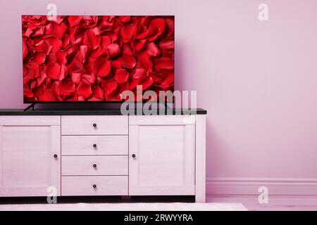 Écran de télévision avec pétales de rose rouge sur commode près du mur rose dans la chambre, espace pour le texte Banque D'Images